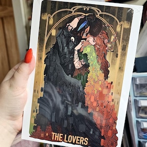 Könnte beinhalten: Ein Tarotkarten-Druck mit zwei sich umarmenden Figuren. Eine Figur ist dunkel mit rabenartigen Flügeln, die andere hat rotes Haar und florale Akzente. Die Karte ist mit goldenen Details umrahmt und die Worte "THE LOVERS" stehen unten.