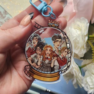 May include: A clear acrylic keyring with a cartoon illustration of five people inside a snow globe. The keyring has a gold base and a blue clasp. The illustration depicts people with various hair colours and outfits.