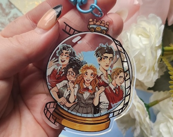 May include: A clear acrylic keychain featuring a cartoon illustration of five people inside a snow globe. The keychain has a gold base and a blue clasp. The illustration depicts people with various hair colors and outfits.