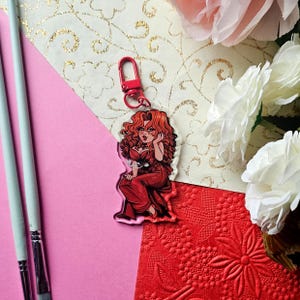 May include: A red-haired woman illustration keyring with a red clasp. The woman is wearing a red dress and is seated with her hand on her chin. The keyring is on a pink and gold patterned background with white flowers.