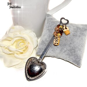 Peut inclure: Une infuseur à thé en forme de cœur argenté avec une longue poignée. L'infuseur est décoré de petites perles colorées et de miniatures de chocolat et de crème glacée. L'infuseur repose sur une surface en tissu gris.