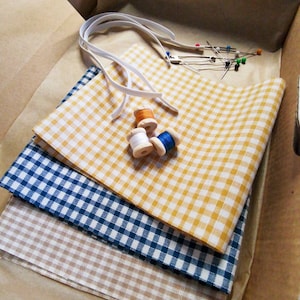 Peut inclure: Quatre morceaux de tissu dans une boîte, dont un vichy jaune et blanc, un vichy bleu et blanc, un vichy beige et blanc et un tissu beige uni. Il y a aussi des bobines de fil et des épingles à coudre.