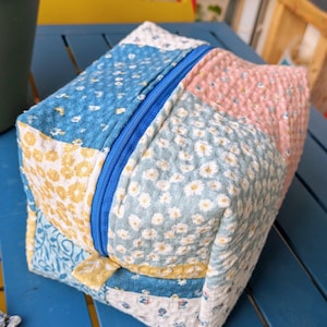 Peut inclure: Une pochette zippée en tissu patchwork bleu, jaune, rose et blanc. La pochette est en tissu matelassé à motif floral.