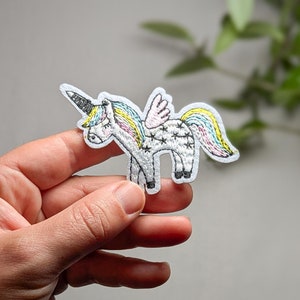 May include: A white unicorn with a rainbow mane and tail, embroidered on a white fabric patch. The unicorn has a black horn and black eyes.