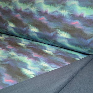 Op de afbeelding: Close-up van stof met een vloeiend, abstract ontwerp. Het patroon bevat tinten blauw, groen en paars met roze accenten. De stof is gevouwen en toont een effen blauwgrijze onderkant. Het geheel doet denken aan een aquarel.
