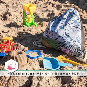 Op de afbeelding: Een blauwe en grijze trekkoordtas met een wit en blauw nautisch patroon. De tas ligt op een zanderig oppervlak met een speelgoedtractor, een kleine blauwe plastic container en een geel en groen zand speelgoed.