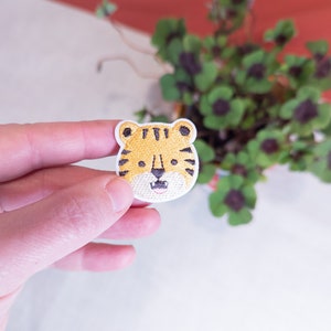 May include: A close-up of a yellow and black embroidered tiger face patch. The patch is held in a hand against a white background.