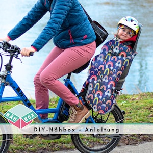 Può includere: Una persona che guida una bicicletta blu con un bambino in un seggiolino per bicicletta a motivi arcobaleno. Il seggiolino ha un design bianco e nero con arcobaleni e nuvole. Il testo "DIY-Nähbox + Anleitung" è visibile sulla bicicletta.