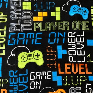 May include: Black fabric with a repeating pattern of video game controllers, pixelated text, and colorful blocks. The text includes "GAME ON", "PLAYER ONE", "LEVEL UP", and "POWER".