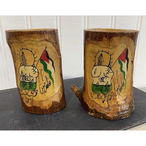 May include: Two wooden cups with a hand-painted design. Each cup features a cartoon figure holding a flag with red, white, green, and black stripes. The figure is wearing green shorts. The cups are made from tree trunks.
