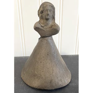 May include: A gray, handcrafted sculpture of a woman's bust atop a conical base. The sculpture has a weathered appearance, with visible texture and subtle color variations. The figure's face is detailed, and the overall design suggests a vintage or antique aesthetic.