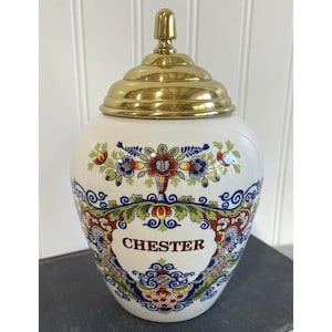 May include: A white ceramic jar with a gold-colored lid and finial. The jar is decorated with colorful floral patterns and the name "CHESTER" in a banner. The jar is likely used for storage.