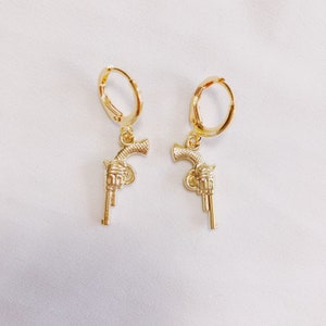 May include: A pair of gold-toned earrings featuring a gun charm. The earrings are hoop style with a simple design.