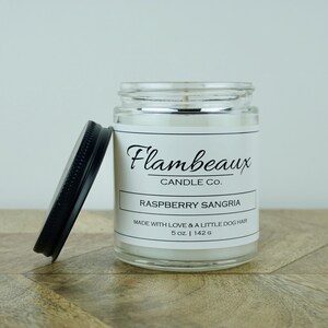 May include: A glass candle jar with a black lid. The label reads "Flambeaux Candle Co. Raspberry Sangria Made with love & a little dog hair 5 oz. | 142 g"