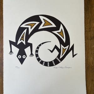 May include: A black and gold lizard design on a white background. The lizard's body forms a curved shape with a patterned tail. The print is numbered 39/100 and signed "Hope Pottery Designs".
