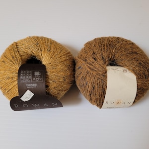 May include: Two skeins of Rowan Felted Tweed yarn. One skein is a golden yellow color with black flecks, and the other is a rich brown. Each skein has a label with the brand name and other information.