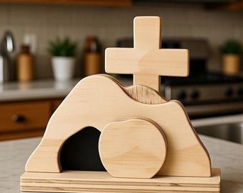 May include: A wooden Easter scene depicting a cross atop a hill with a tomb. The tomb has a dark interior and a circular stone in front. The scene is made of light-colored wood and sits on a rectangular base.