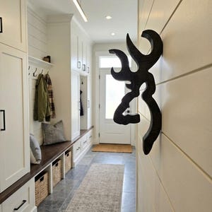May include: A dark brown, wooden wall-mounted deer head silhouette featuring a stylized, abstract antler design. The object is mounted on a white horizontal shiplap wall, positioned in a hallway with white cabinetry and a light-colored runner rug.