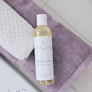 Puede incluir: Una botella transparente de baño de burbujas de lavanda con una etiqueta blanca que dice "Forest Field Flowers Bubbles & Bliss Lavender Bubble Bath Bain Moussant à la lavande 250 ml". La botella está sobre una toalla morada.