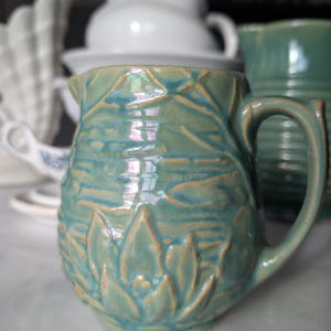 May include: A turquoise ceramic pitcher with a detailed floral design. The pitcher has a curved handle and a spout for pouring. Other white and turquoise pitchers are in the background.