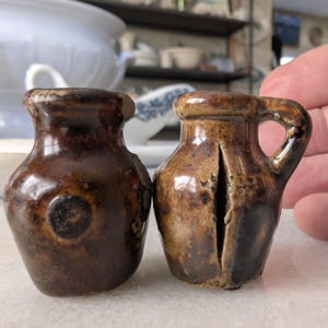 May include: Two small, antique brown ceramic jugs. The jug on the left is a dark brown with a circular design. The jug on the right is a lighter brown with a handle and a vertical slit. Both jugs have a glossy finish.