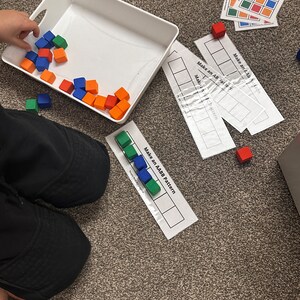 Op de afbeelding: Een kind speelt met kleurrijke blokken. De blokken zijn rood, blauw, groen en oranje. Het kind gebruikt de blokken om patronen te maken op een wit vel papier. Het papier heeft de tekst "Make an AABB Pattern" erop gedrukt.