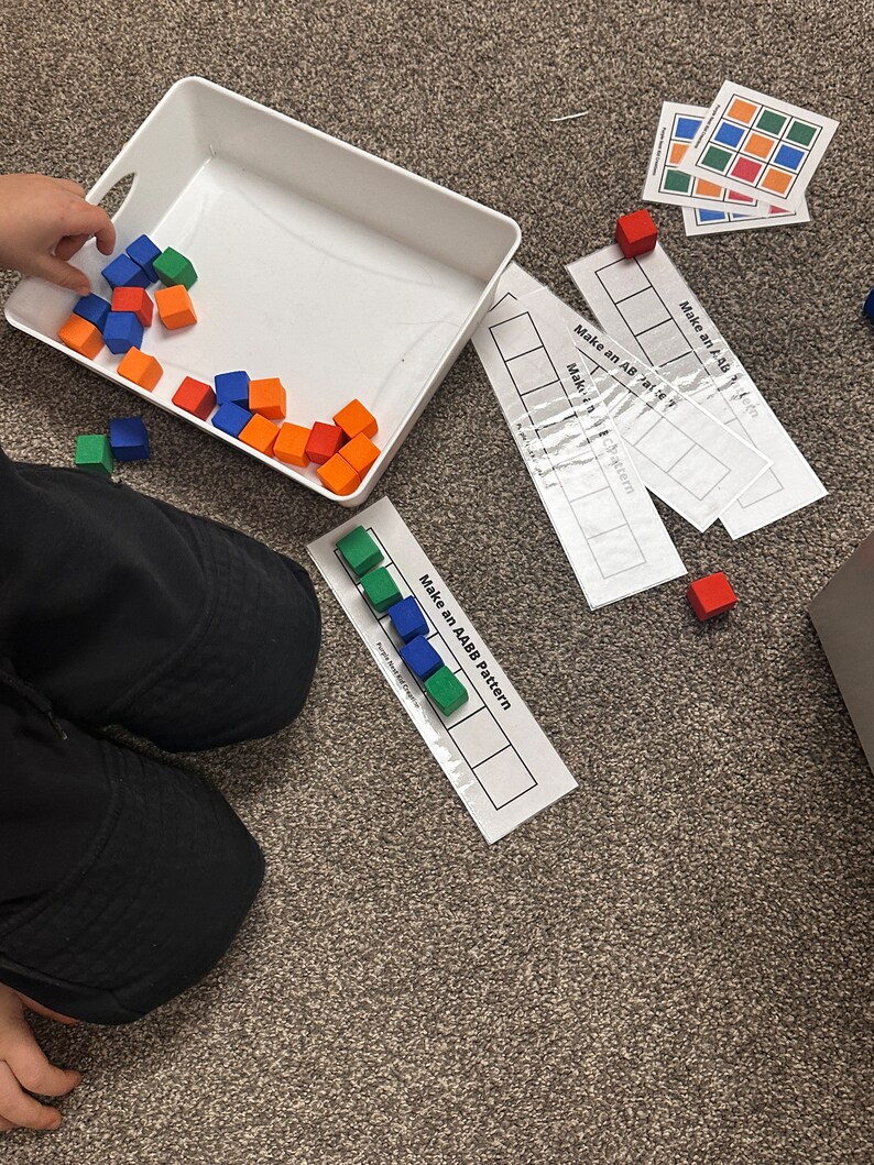 Op de afbeelding: Een kind speelt met kleurrijke blokken. De blokken zijn rood, blauw, groen en oranje. Het kind gebruikt de blokken om patronen te maken op een wit vel papier. Het papier heeft de tekst "Make an AABB Pattern" erop gedrukt.