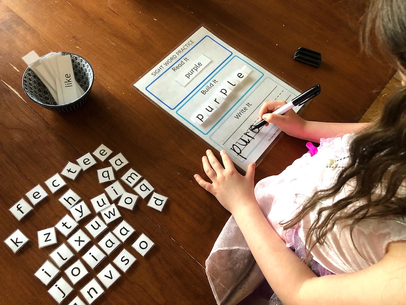 May include: A child is learning to spell the word "purple" using letter tiles. The word is written on a white sheet of paper with the words "Read It", "Build It", and "Write It" above it. The child is using a black marker to write the word.