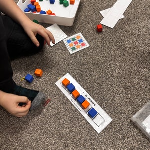 Op de afbeelding: Een kind speelt met een set kleurrijke houten blokken. De blokken zijn gerangschikt in een patroon op een witte kaart met de woorden "AB Pattern" erop gedrukt. Het kind gebruikt de blokken om een patroon te maken.