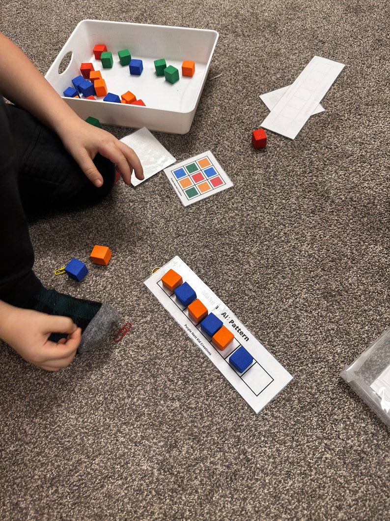 Op de afbeelding: Een kind speelt met een set kleurrijke houten blokken. De blokken zijn gerangschikt in een patroon op een witte kaart met de woorden "AB Pattern" erop gedrukt. Het kind gebruikt de blokken om een patroon te maken.