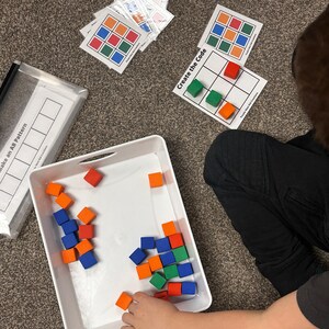 Op de afbeelding: Een kind speelt een spel met kleurrijke schuimblokken. De blokken bevinden zich in een witte bak. Het kind matcht de blokken met een raster op een kaart waarop "Maak de code" staat.