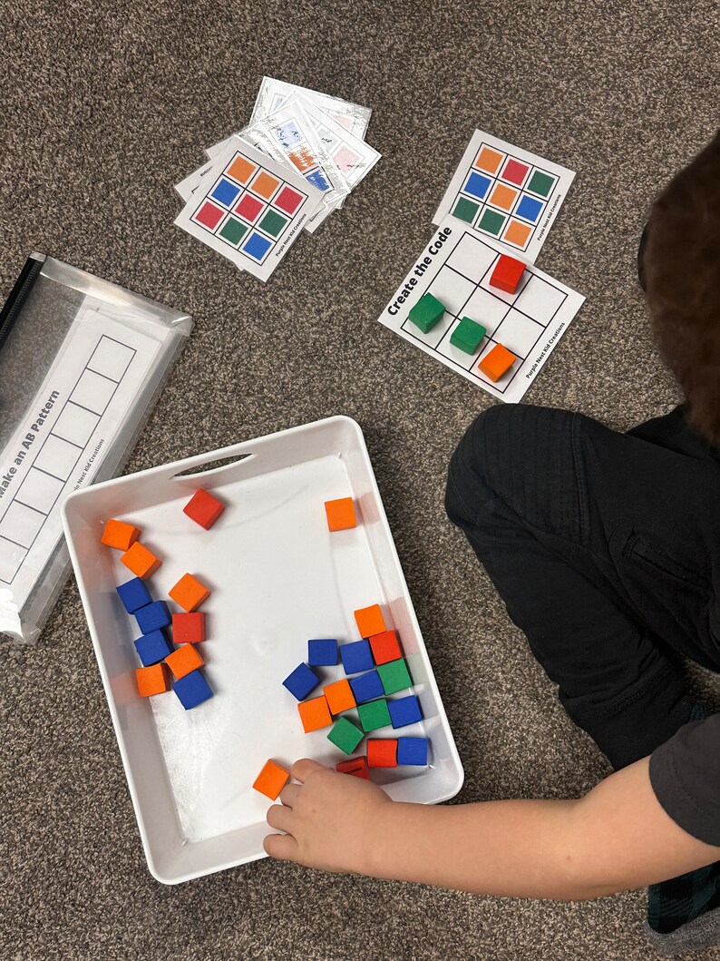 Op de afbeelding: Een kind speelt een spel met kleurrijke schuimblokken. De blokken bevinden zich in een witte bak. Het kind matcht de blokken met een raster op een kaart waarop "Maak de code" staat.