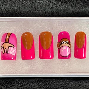 May include: Five pink artificial fingernails with different designs. One nail has a brown tip and a cartoon monkey blowing a bubble. Another nail has a brown tip and a cartoon arm reaching out. The remaining three nails have brown tips.
