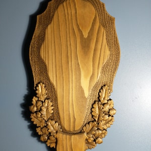 Könnte beinhalten: Eine Holzplakette mit einem geschnitzten Design aus Eichenblättern und Eicheln. Die Plakette ist braun und hat eine strukturierte Oberfläche.