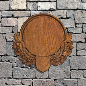 May include: A wooden plaque with a circular center and a decorative oak leaf and acorn border. The plaque is mounted on a gray stone wall. The wood has a natural grain and a warm brown color.