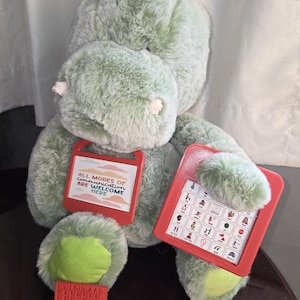 May include: A soft, green dinosaur toy with a red communication board and a red keychain. The dinosaur has a fuzzy texture and a green belly. The communication board displays various symbols. The keychain says "WELCOME".