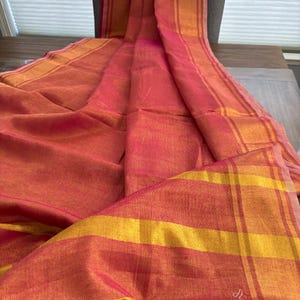 May include: A long, rectangular textile piece featuring a vibrant blend of hot pink and golden yellow hues. The fabric displays a prominent striped pattern with alternating wide bands of pink and gold, creating a textured, woven appearance throughout.