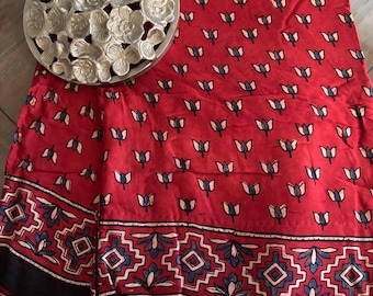 May include: A red fabric featuring a repeating pattern of small dark blue and light pink tulip-like flowers. The bottom edge displays a wide border with a geometric diamond motif in red, blue, and pink. A silver circular floral sculpture sits on top.
