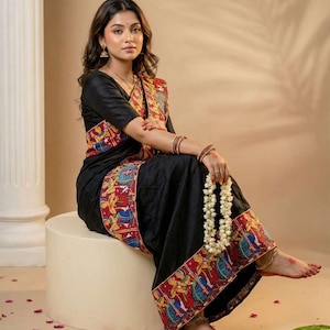 May include: A black saree featuring a wide border with colorful hand-painted illustrations of figures in red, blue, and yellow. The garment includes a matching black short-sleeved blouse and is draped to showcase the detailed folk art patterns.