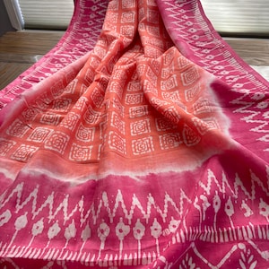 May include: A vibrant textile featuring a repeating square pattern in shades of coral and white, transitioning to a bright pink with a geometric design. The fabric is draped, showcasing its texture and color variations.