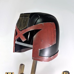 Op de afbeelding: Een zwart en rode helm met een opvallend goudkleurig embleem op het voorhoofd. De helm heeft een verweerde afwerking met zichtbare krassen en een uitgesproken hoekig ontwerp, inclusief een brede vizieropening en een kruisvormig detail.