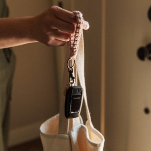 May include: A pink crocheted key chain with a black key fob attached to a white tote bag.