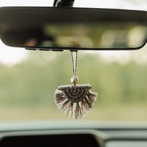 Op de afbeelding: Een beige macramé autoluchtverfrisser die hangt aan de achteruitkijkspiegel van een auto. De luchtverfrisser heeft de vorm van een halve cirkel met kwastjes aan de onderkant.