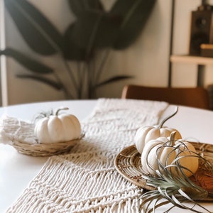 May include: White pumpkins on a woven table runner with a green plant in a wicker basket. The table runner is made of a thick, knotted rope and has a fringe.