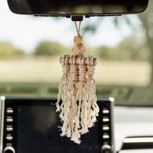 Pode incluir: Um aromatizador de carro de macramé branco com contas de madeira penduradas no espelho retrovisor de um carro.