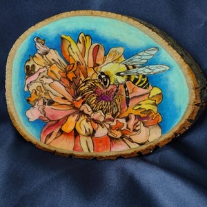 May include: A hand-painted wooden slice featuring a detailed illustration of a bee pollinating a flower. The artwork showcases a yellow and black bee with transparent wings, set against a blue sky and a vibrant orange and pink flower.