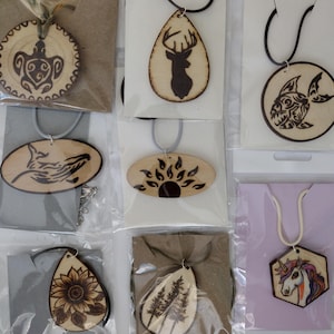 May include: A collection of wooden pendant necklaces with various designs, including a turtle, deer, fish, wolf, sun, sunflower, trees, and a unicorn. Each pendant is attached to a cord or string, and packaged in clear plastic.