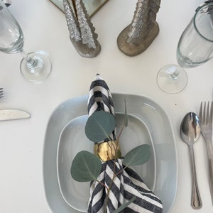 May include: A white square plate with a smaller white square plate inside. A black and white striped napkin is placed on the smaller plate with a gold napkin ring and a sprig of eucalyptus leaves.