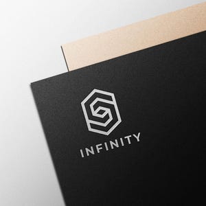Op de afbeelding: Een zwarte visitekaart met het woord "INFINITY" in zilver, met daarboven een geometrisch logo in zilver. Op de achtergrond is gedeeltelijk een beige kaart zichtbaar. Het logo en de tekst zijn in een schreefloze lettertype.