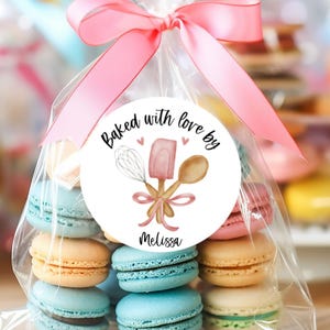 May include: A clear cellophane bag tied with a pink ribbon, filled with colorful macarons. A round sticker on the bag reads "Baked with love by Melissa" with baking tools. The macarons are various colors, including blue, orange, and pink.